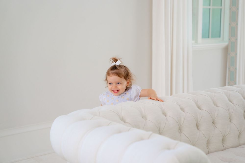 Young child plays joyfully on a tufted white sofa in a bright, minimalist room with light walls.