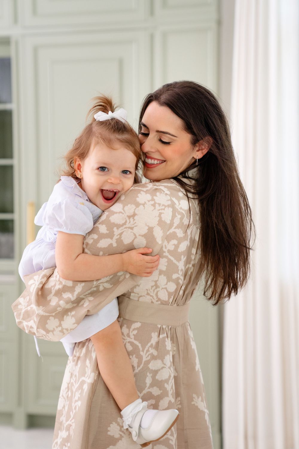 Woman in a floral dress embraces and shares a joyful moment with a laughing toddler in a bright white room.