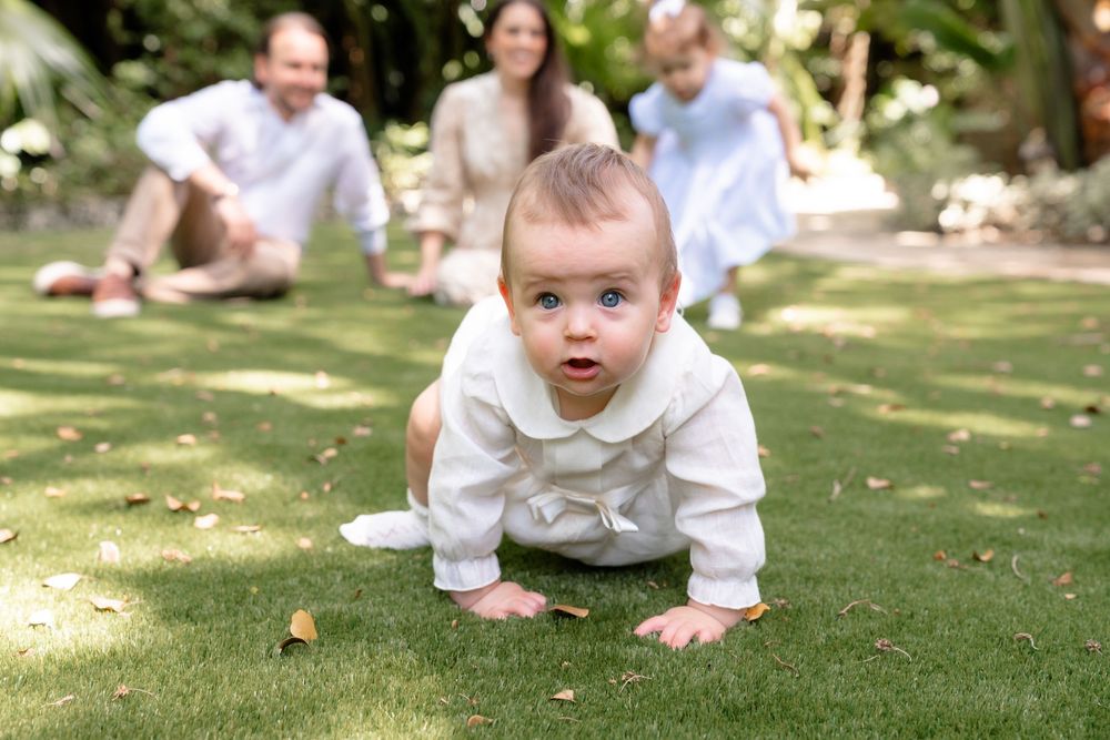 A small child in white clothing crawls on a sunny lawn while others gather in the background on a summer day.
