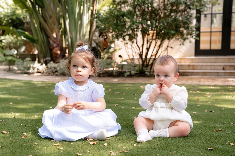 A sequence of connected images showing toddlers in white outfits sitting on grass and enjoying outdoor playtime.