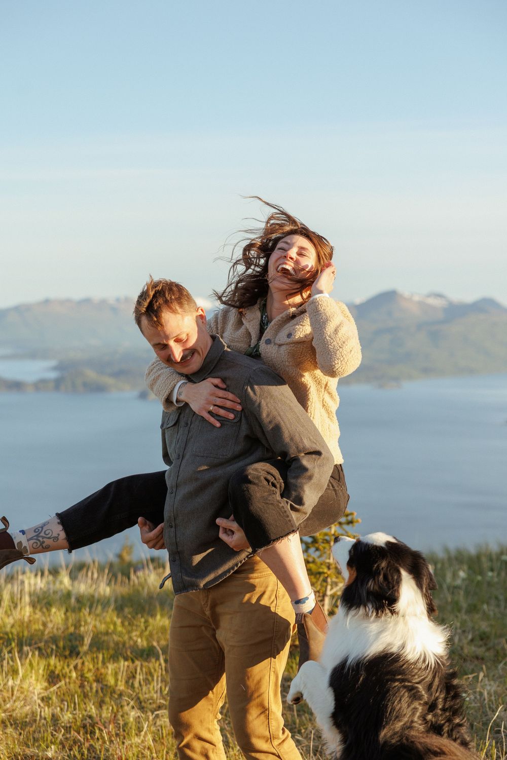 Playful couple spins and dances on a hilltop overlooking a bay with their dog watching.