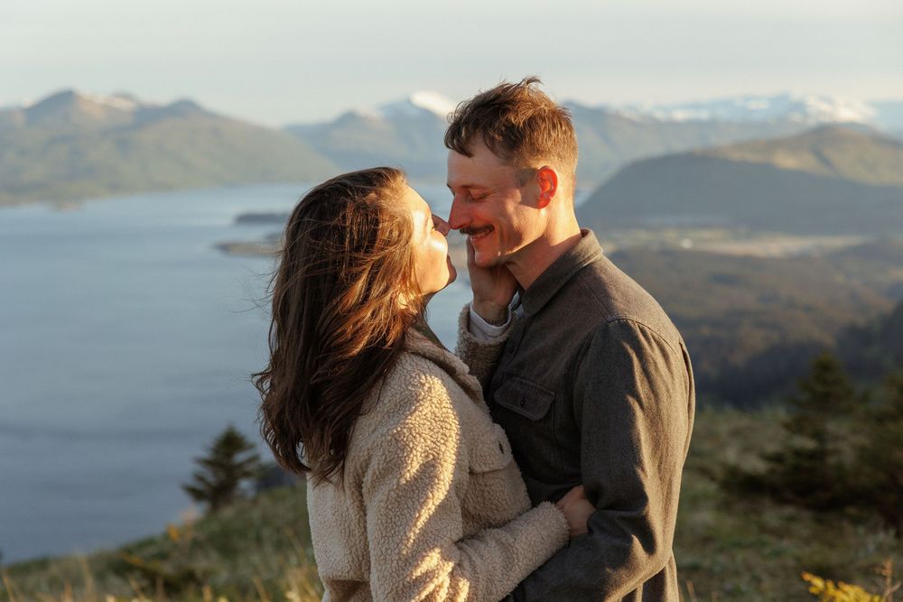 Couple embraces on a scenic mountain overlook with views of a lake and distant peaks at sunset.