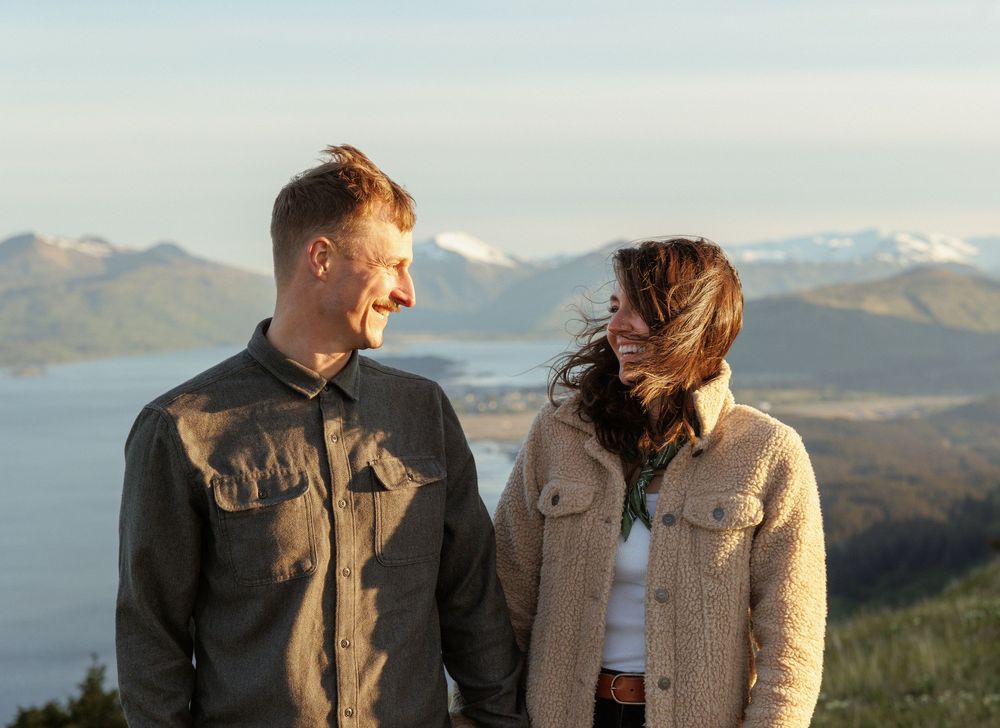 A couple in denim jackets shares a candid moment against a backdrop of snow-capped mountains and rolling hills.