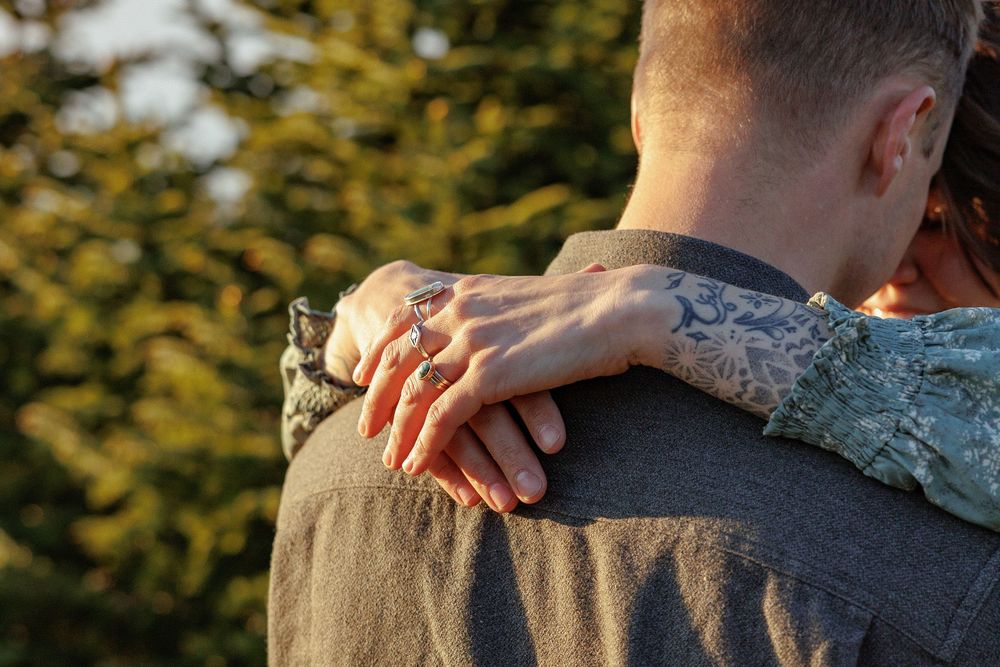 Two people sharing a tender embrace outdoors with one person's tattooed arm visible against golden sunlight.