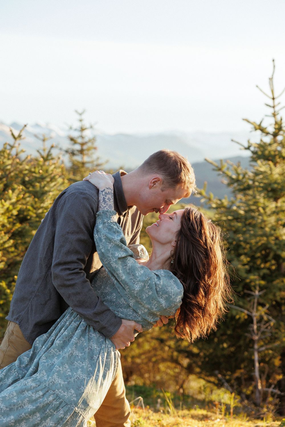 Romantic embrace on a mountain summit with evergreen trees in the background during sunset.