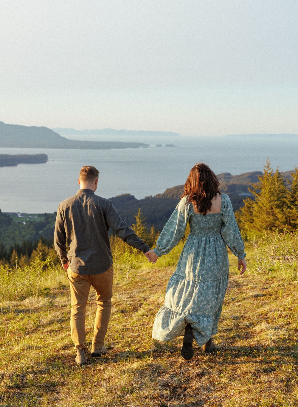 A couple holds hands while walking on a hilltop trail overlooking a serene bay and mountain landscape.