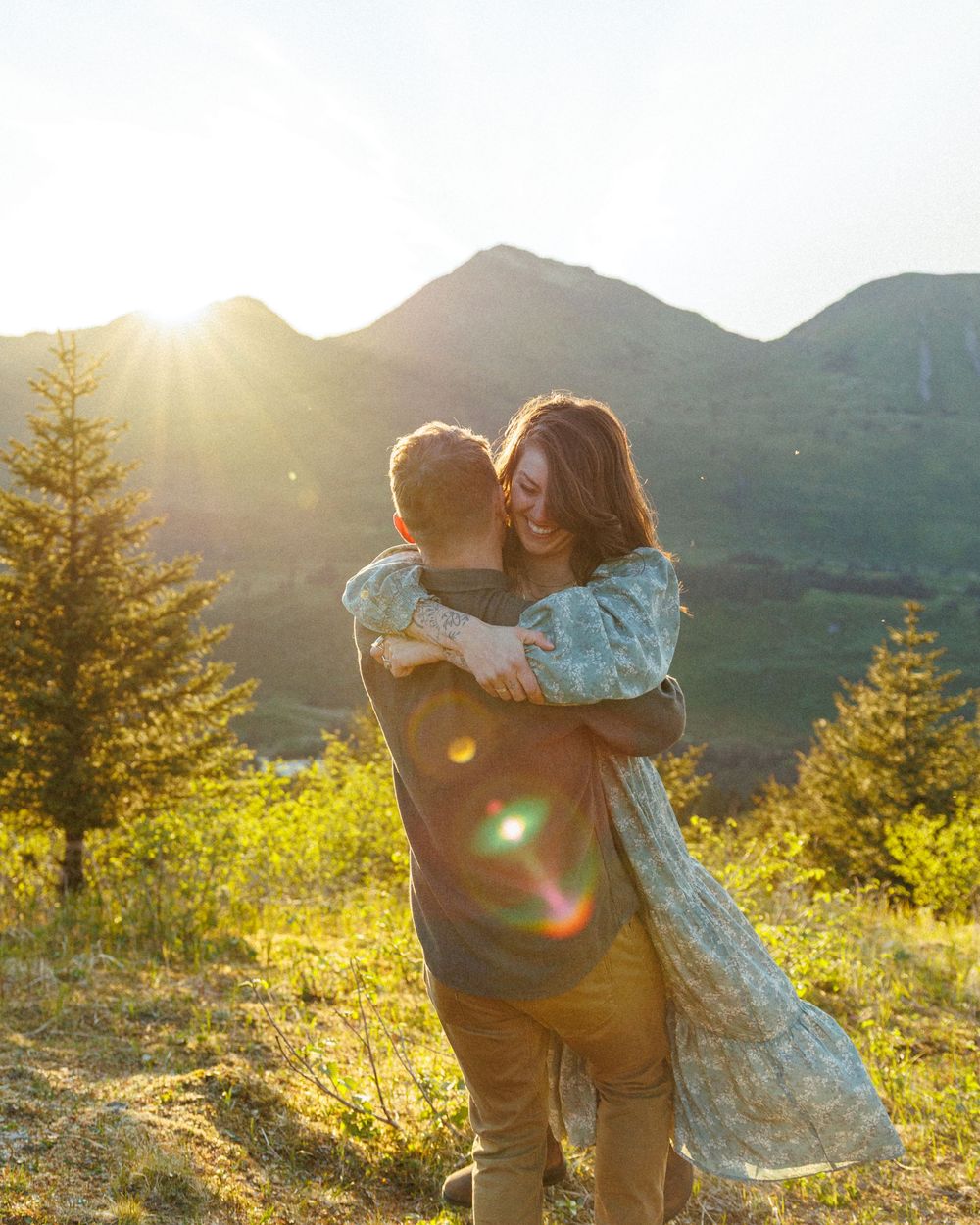 A couple embraces in a mountain meadow at sunset with evergreen trees and sun flares in the background.