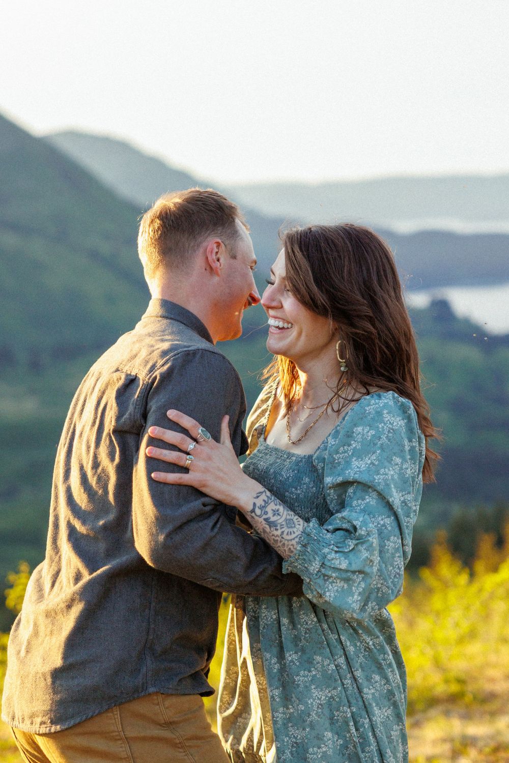 A couple embraces on a mountaintop during golden hour, dressed in blue and denim against a mountainous backdrop.