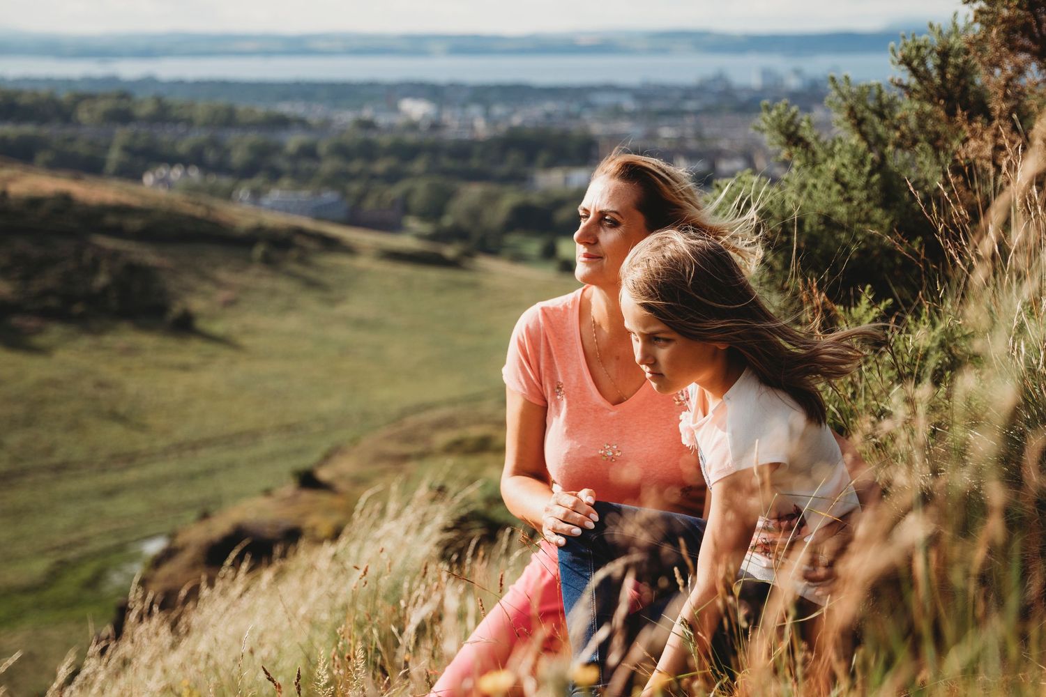 Two people sitting on a grassy hillside overlooking a scenic valley view at sunset.
