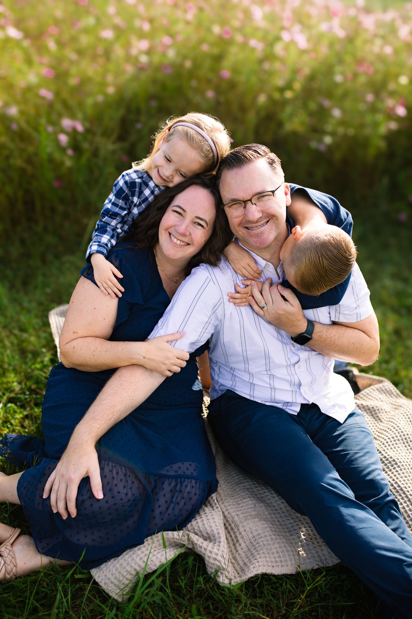 A playful couple with their two kids relaxes together on a blanket outdoors with pink wildflowers and golden sunlight in the background.