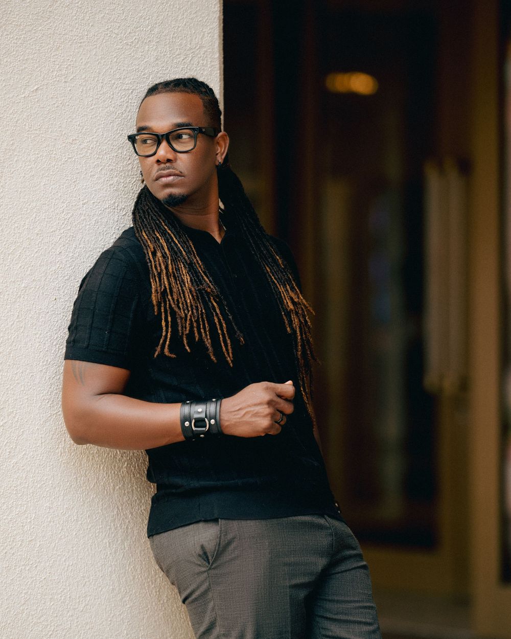 Person with dreadlocks and glasses wearing a black t-shirt and gray pants poses against a light colored wall.