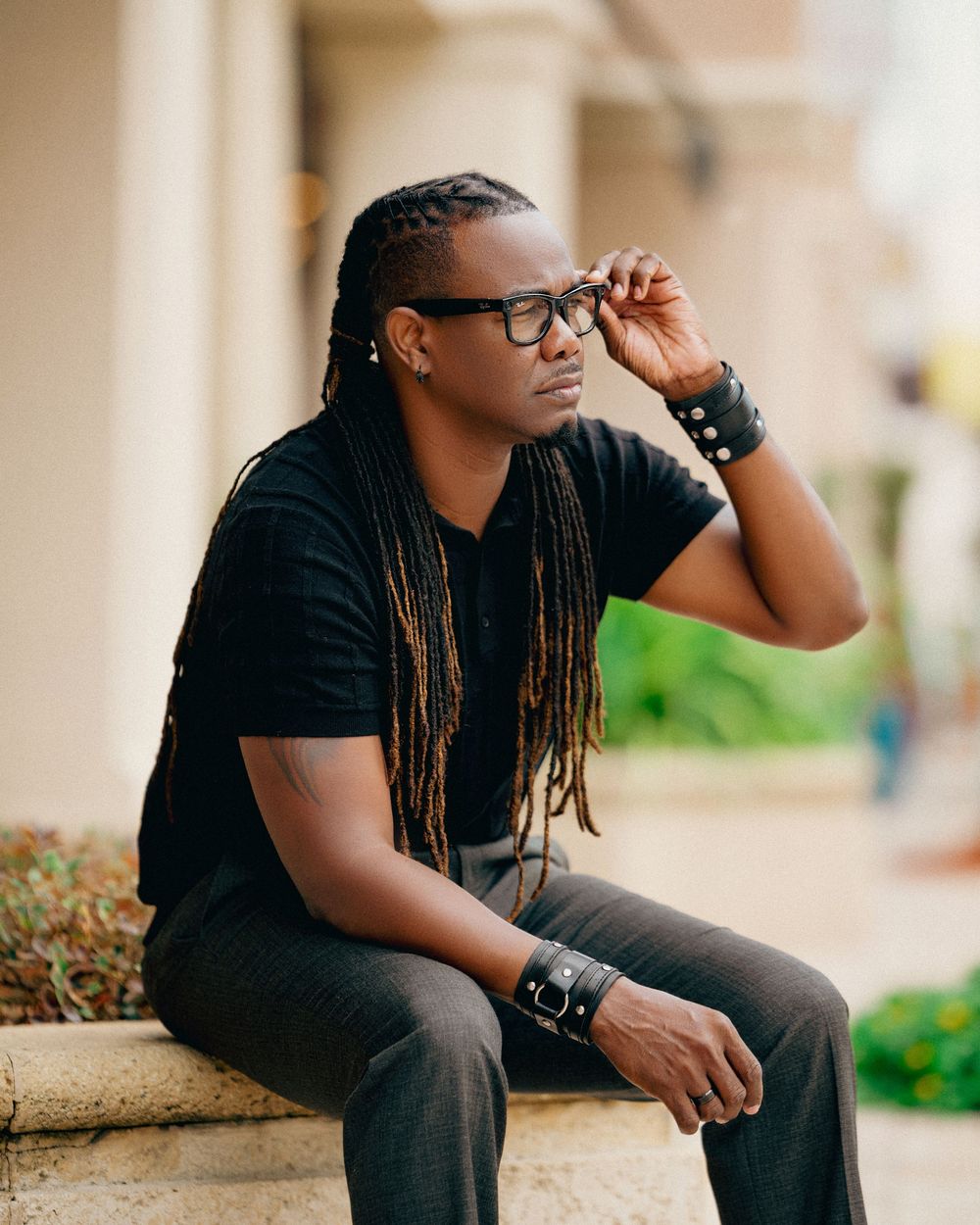 Person wearing glasses and black t-shirt sitting thoughtfully on stone steps with long braided hair and casual style.