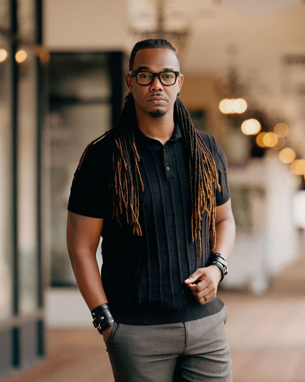 Stylish person in black polo shirt and glasses poses indoors with long dreadlocks and leather accessories.