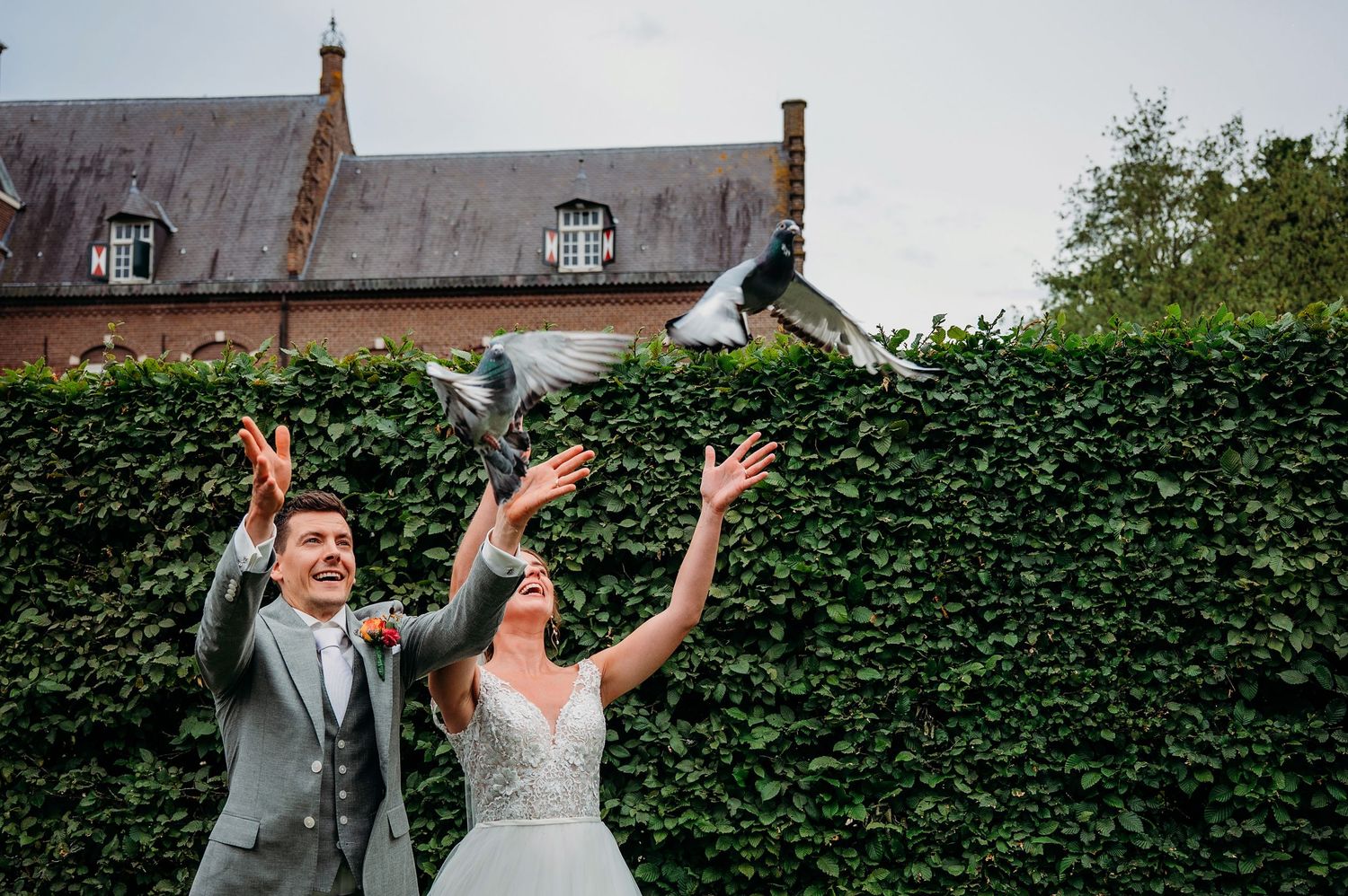 A bride and groom celebrate joyfully in front of a tall hedge wall with a rustic house in the background.