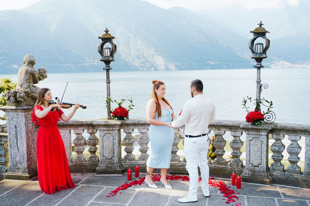 A scenic lakeside terrace with ornate stone balustrades and Mediterranean mountains in the background on a sunny day.