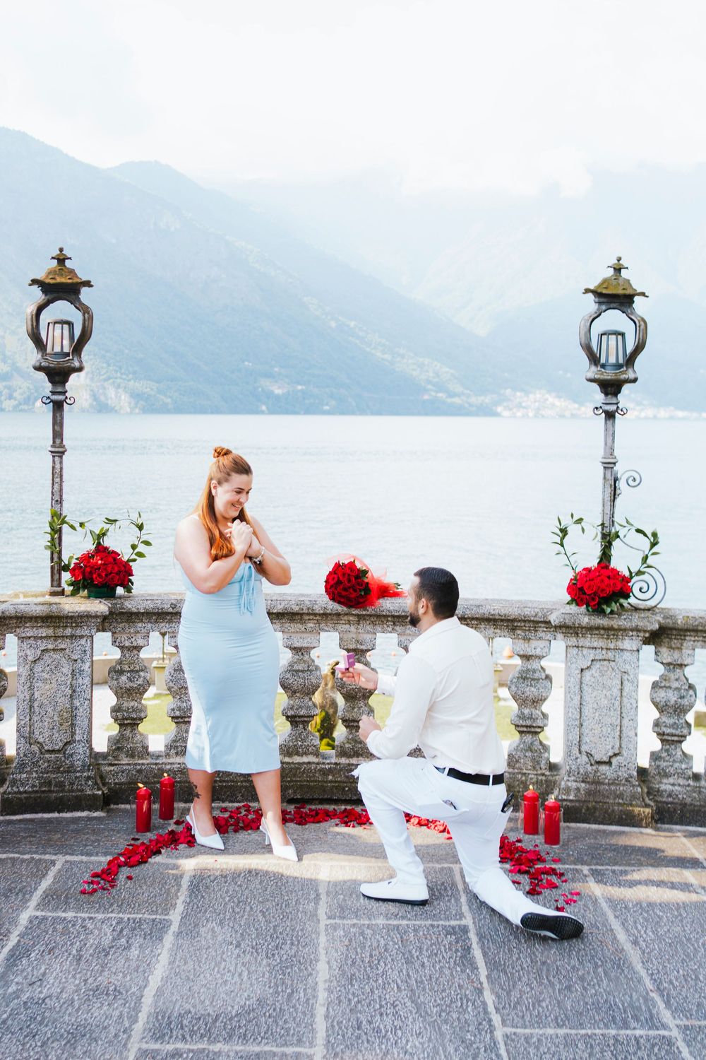 A proposal scene unfolds on a grand stone balcony overlooking Lake Como in Italy with red roses decorating the setting.