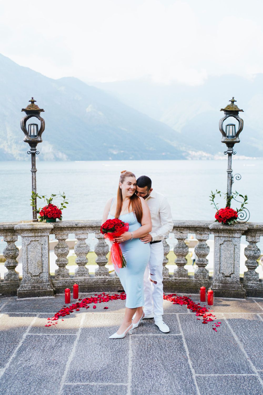Romantic couple embraces on ornate stone balcony with red rose petals and vintage lamps overlooking Lake Como, Italy.
