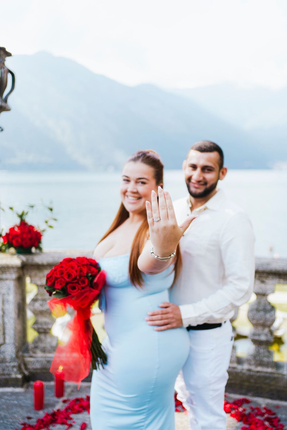 A couple poses for an engagement photo by a scenic lake view with red roses and decorations.