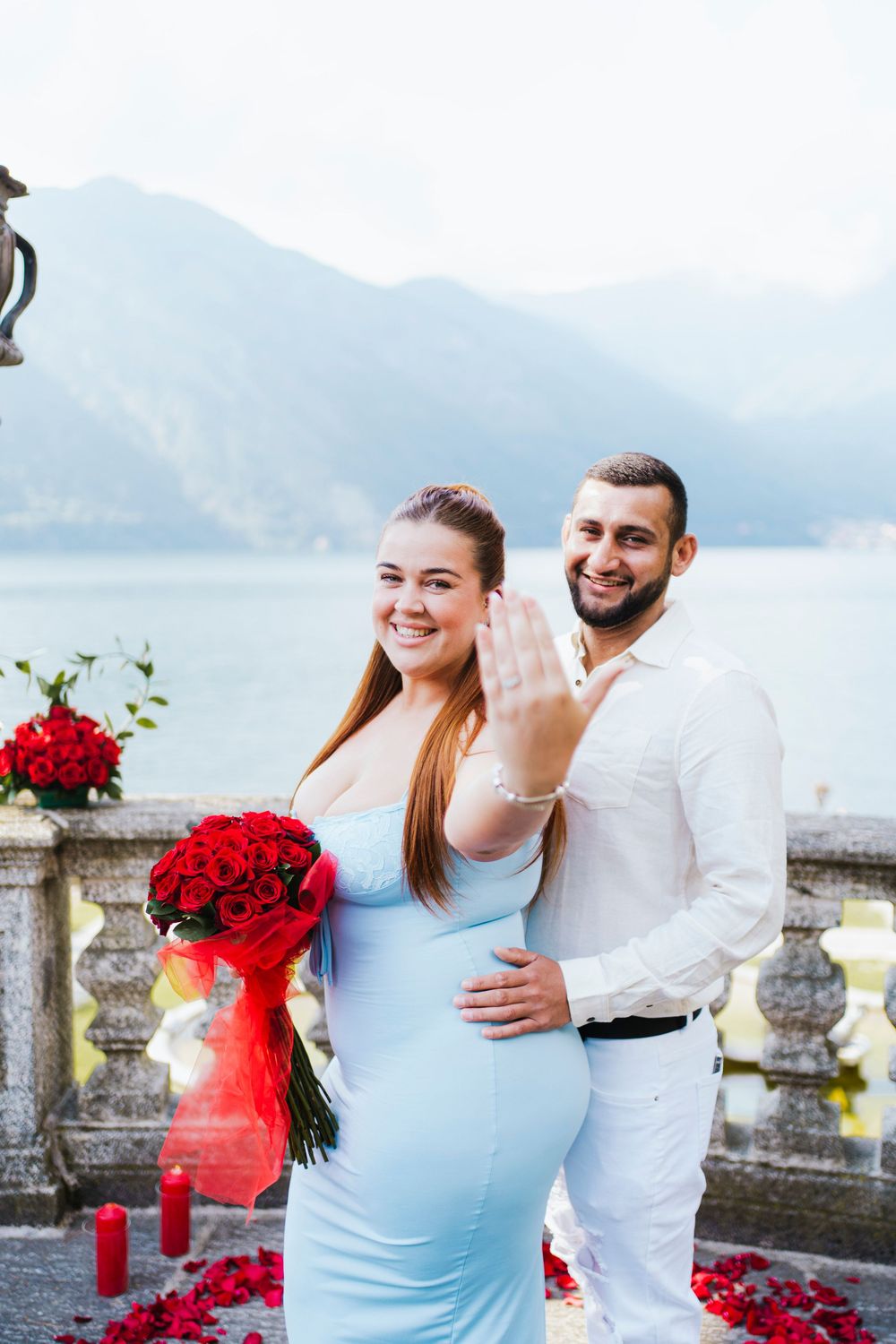 Couple in white attire celebrating romantic moment with red roses on stone balcony overlooking scenic mountain lake.