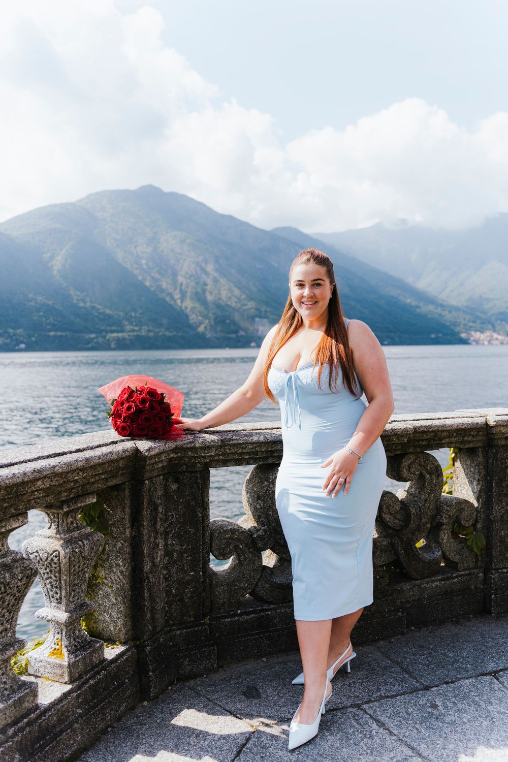 Woman in elegant white dress poses with red bouquet against scenic Lake Como and mountain backdrop in Italy.