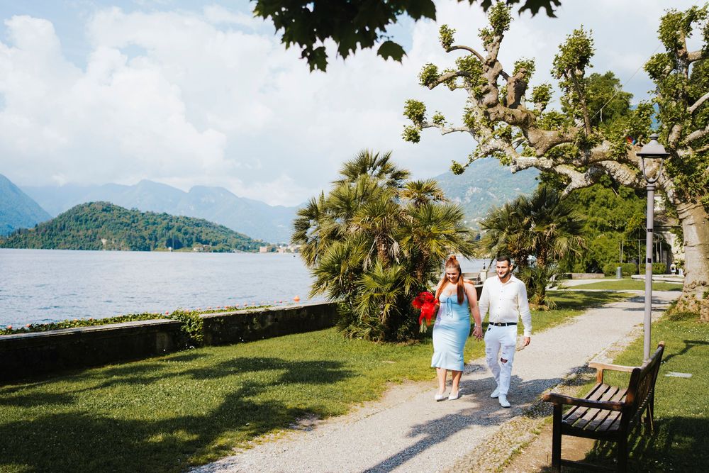 People stroll along a scenic lakeside path with mountains and palm trees on a sunny day in Lake Como, Italy.