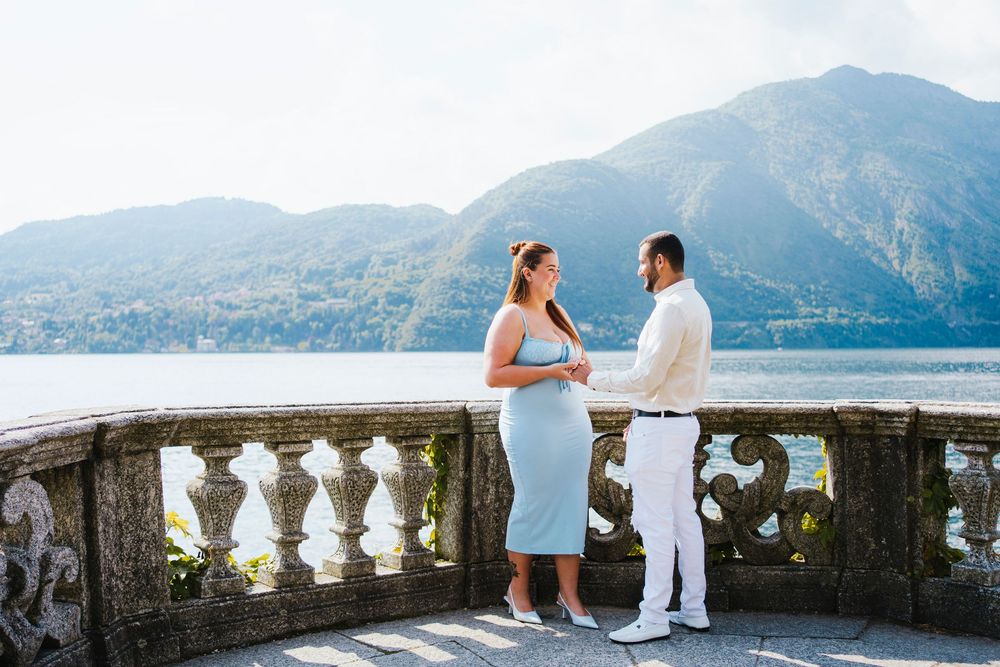 A couple stands on a stone balcony overlooking Lake Como in Italy with mountains in the background.
