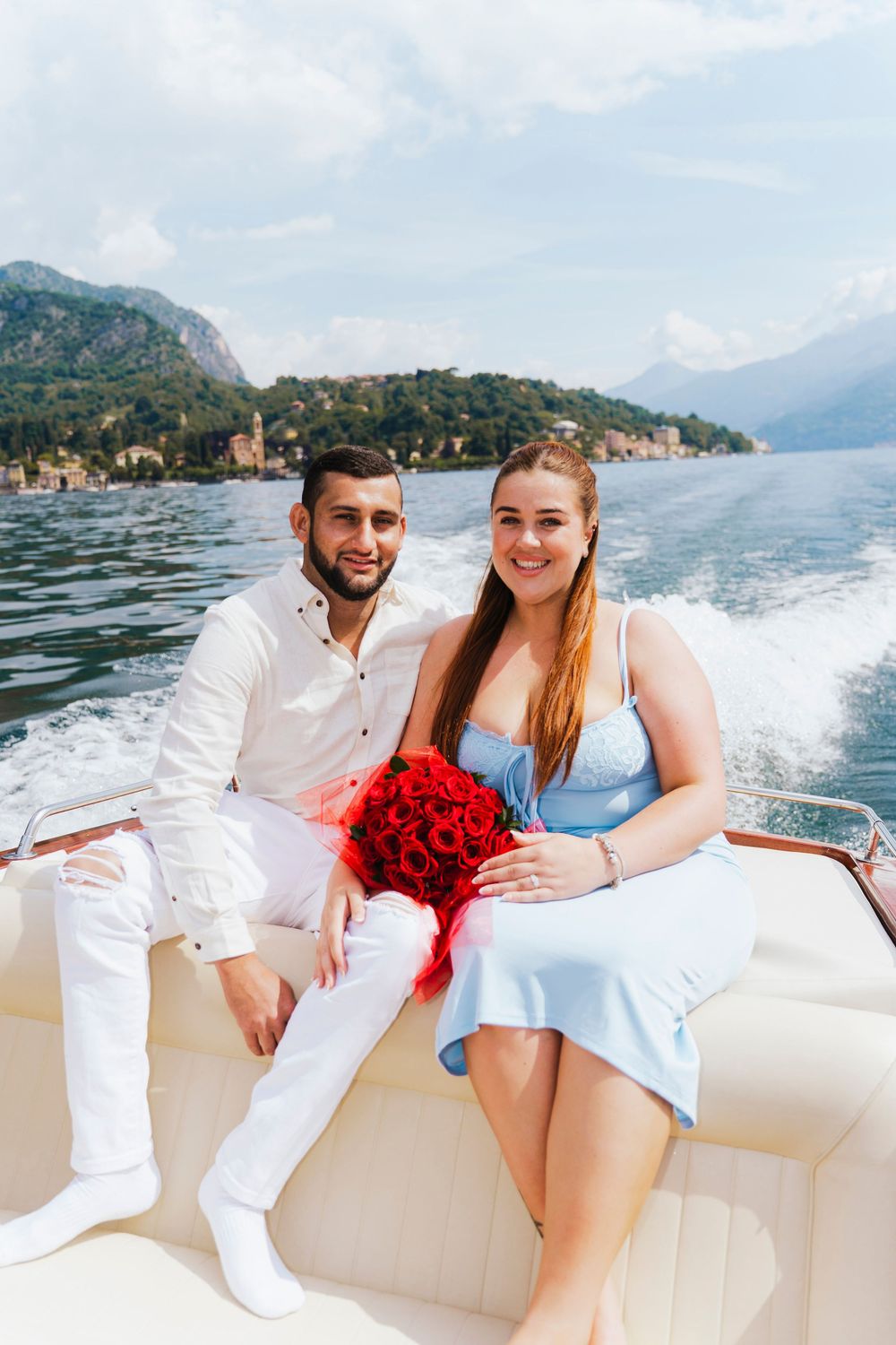 Couple sitting on a boat with red roses against scenic Lake Como backdrop with mountains and waterfront buildings.