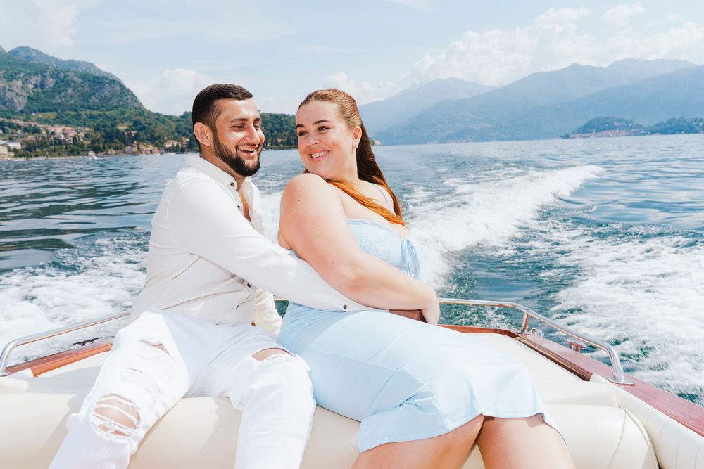 A couple dressed in white embraces while enjoying a romantic boat ride along a scenic mountainous coastline.