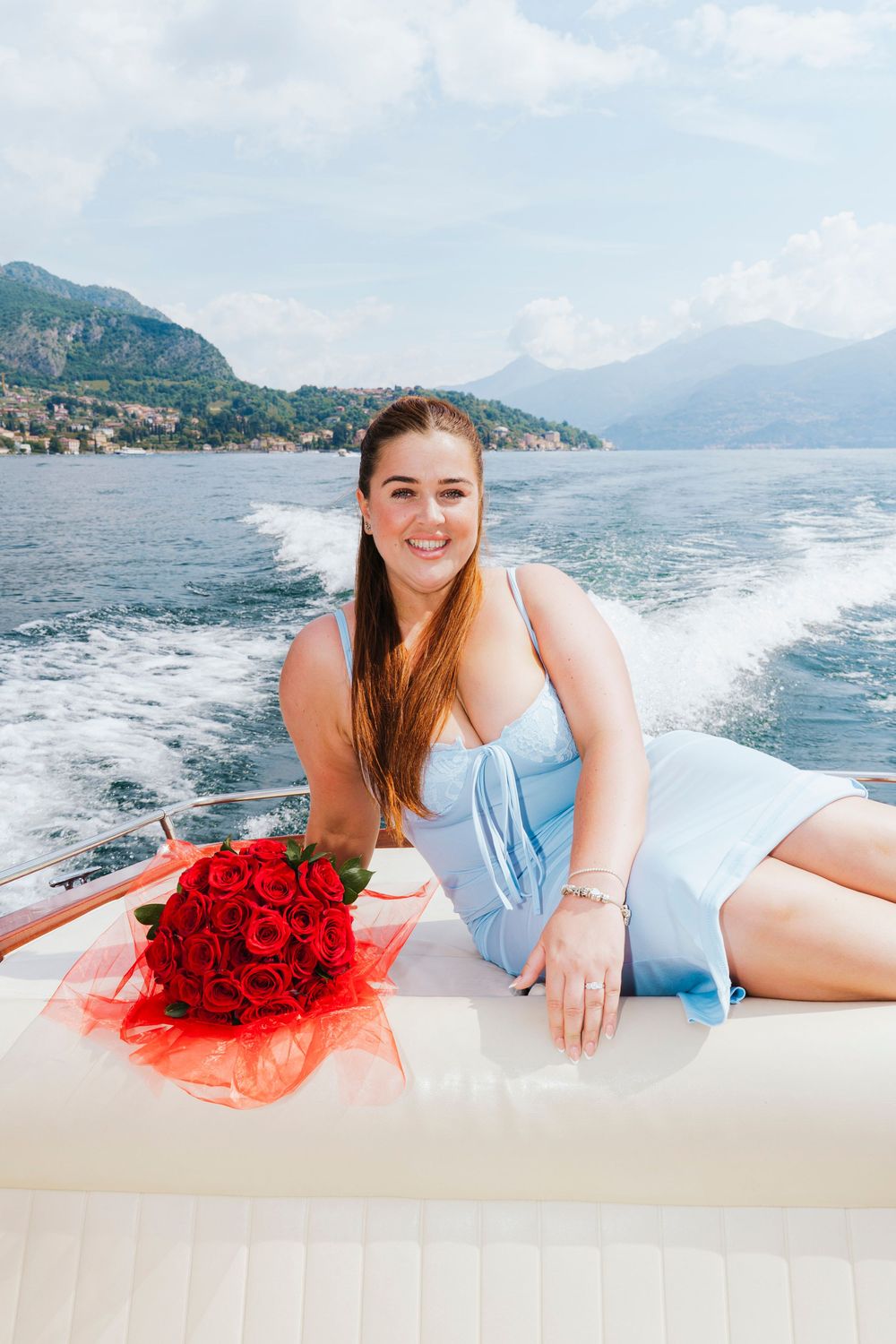 Someone in a light blue dress relaxes on a boat with red roses while cruising past mountains on a scenic lake.