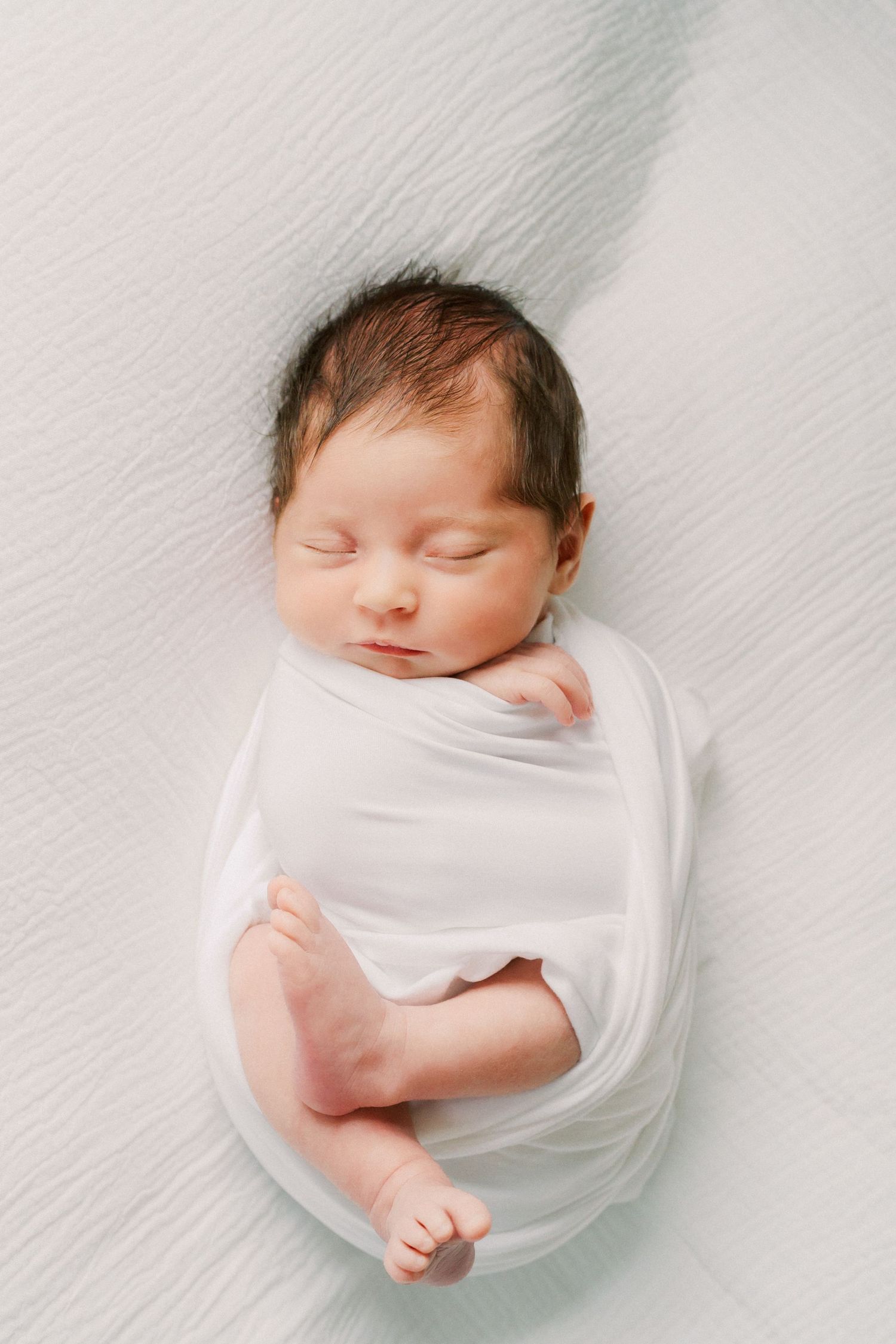A sleeping newborn baby swaddled in soft white fabric lies peacefully against a light background.