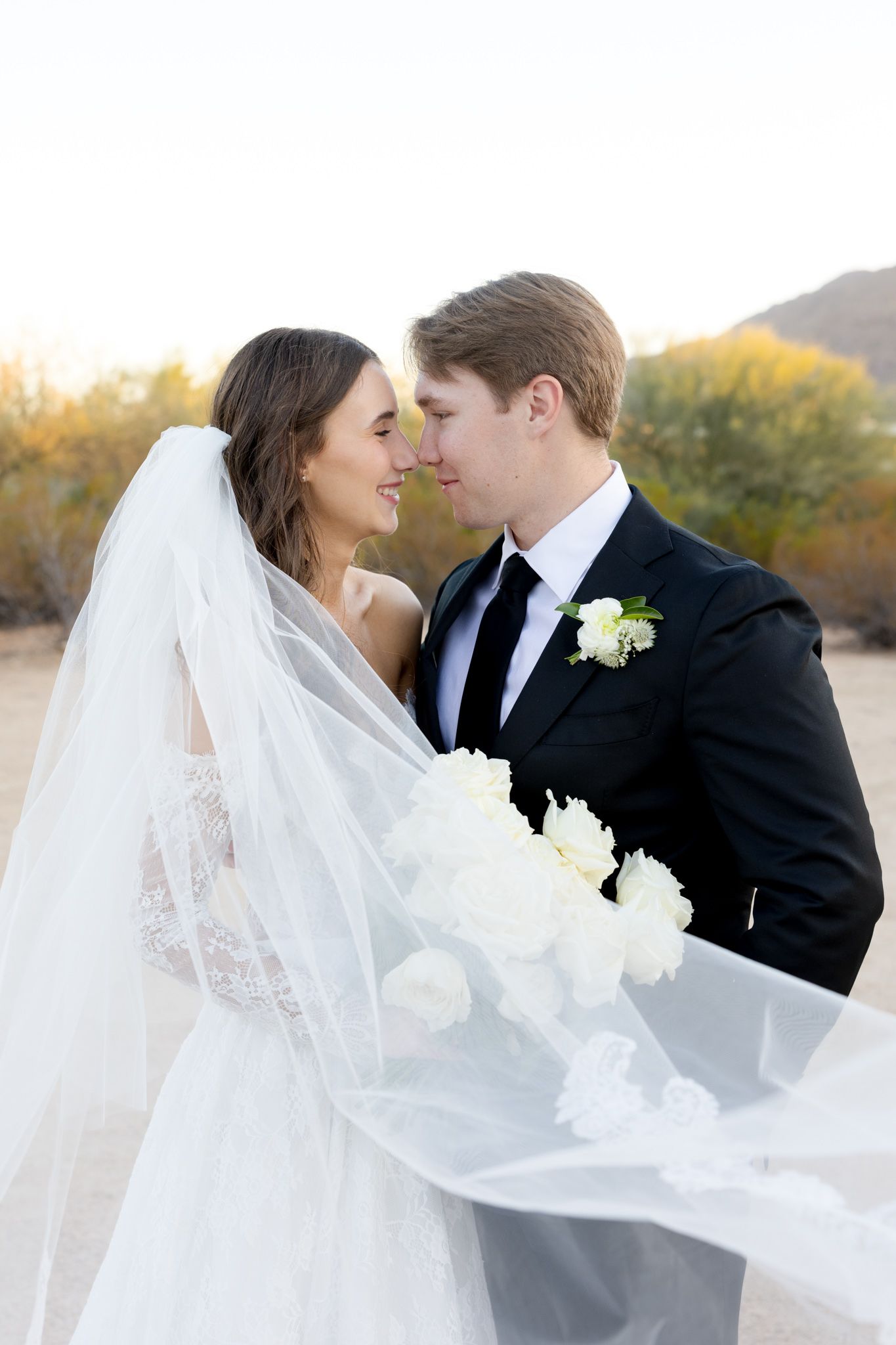 A romantic wedding portrait in a desert landscape at sunset with flowing white veil and white rose boutonniere.