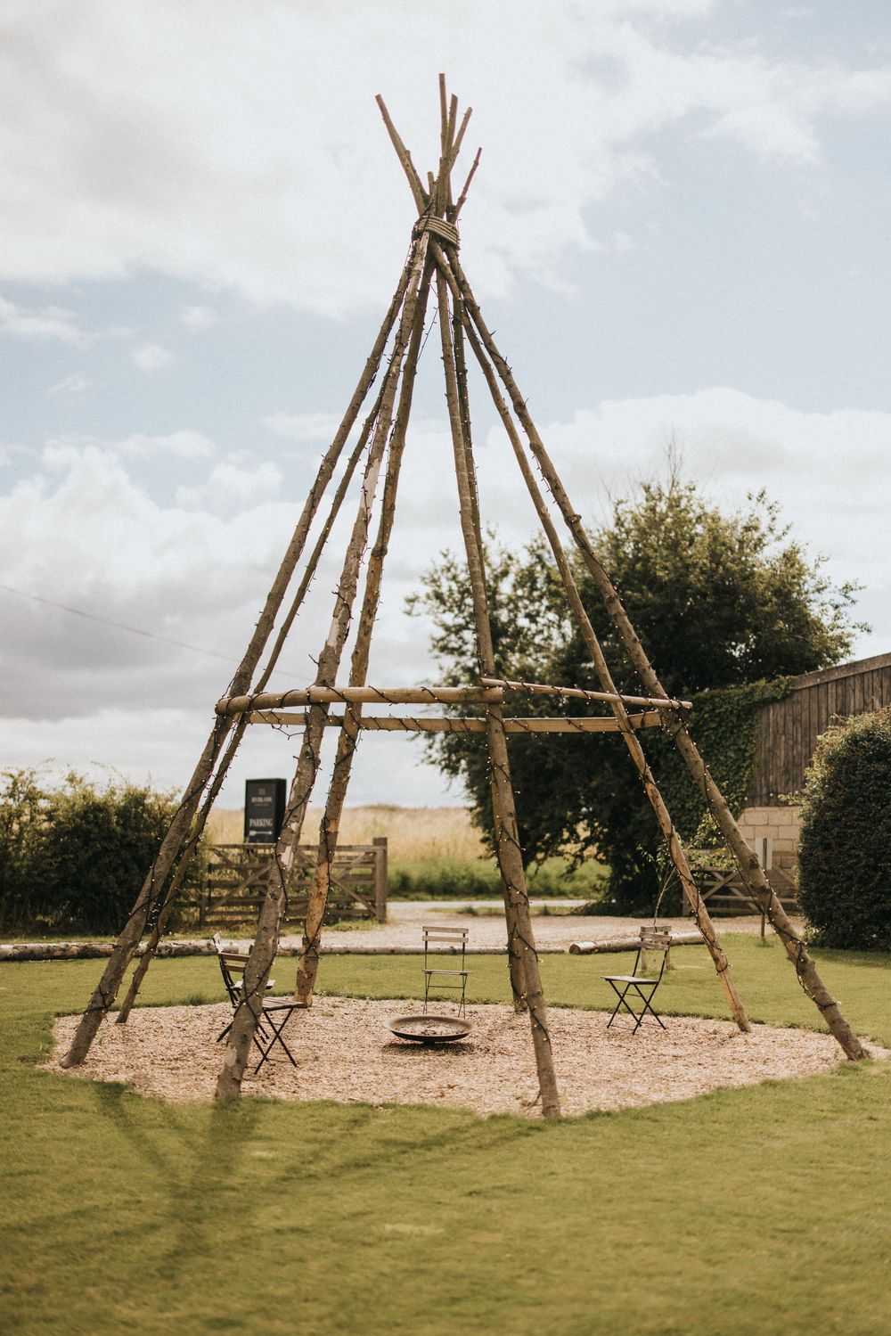 Rustic wooden tipi-style structure with tall poles creates a unique outdoor gathering space over a circular mulch area.