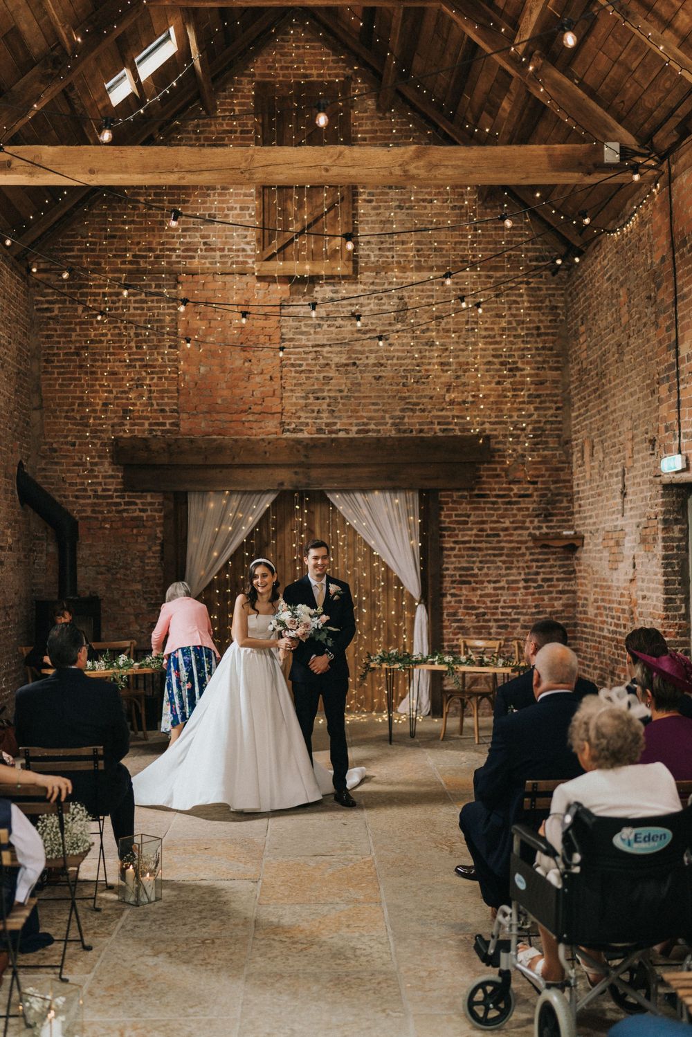 Wedding ceremony setup in an industrial brick venue with string lights and wooden ceiling beams adding warmth.