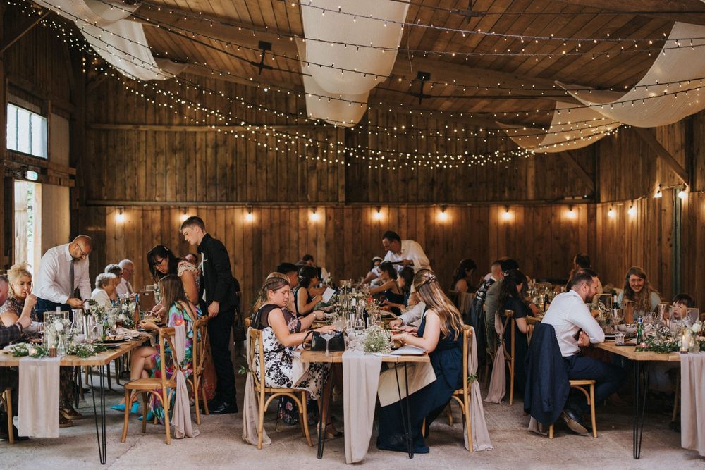 A panoramic view of a rustic barn reception venue with string lights and guests seated at long dining tables.