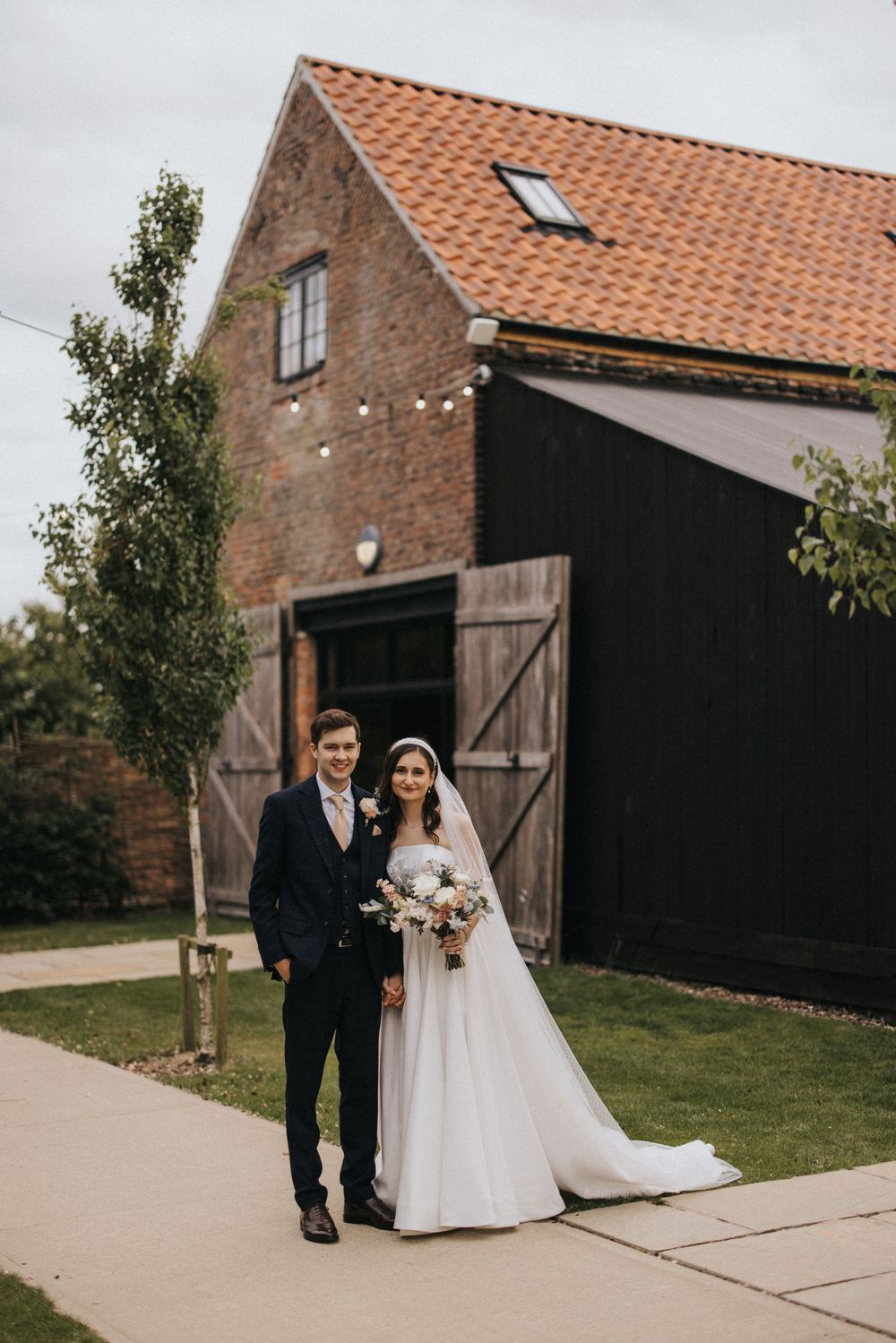 Couple poses together at rustic barn venue with classic red brick walls and terracotta roof tiles.