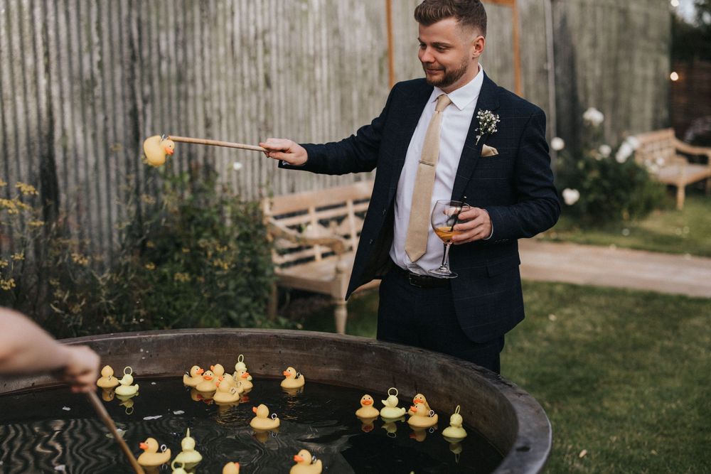 A guest in a navy suit points at floating candles arranged in a rustic outdoor water basin at dusk.