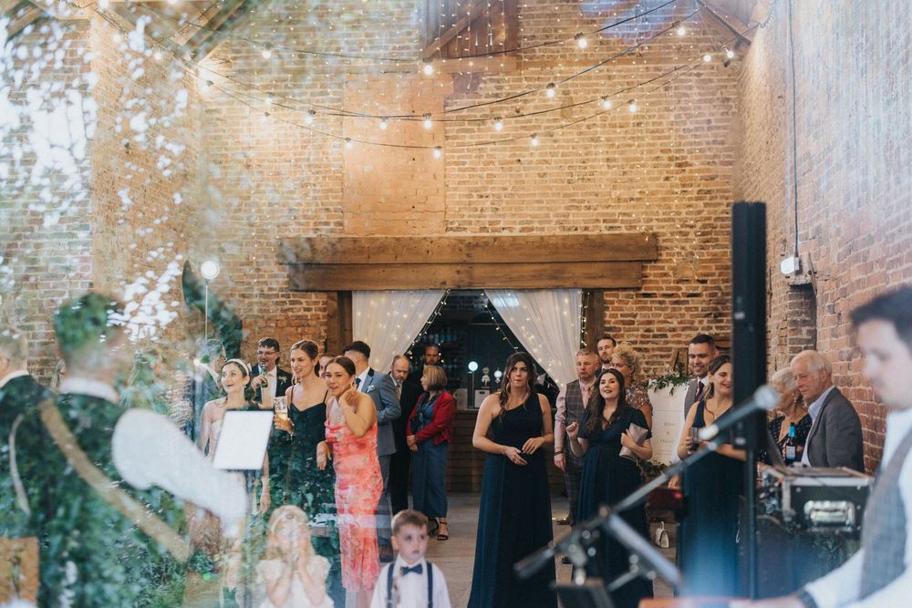 Wedding guests enjoy a reception celebration inside a rustic brick venue with arched windows and string lights.