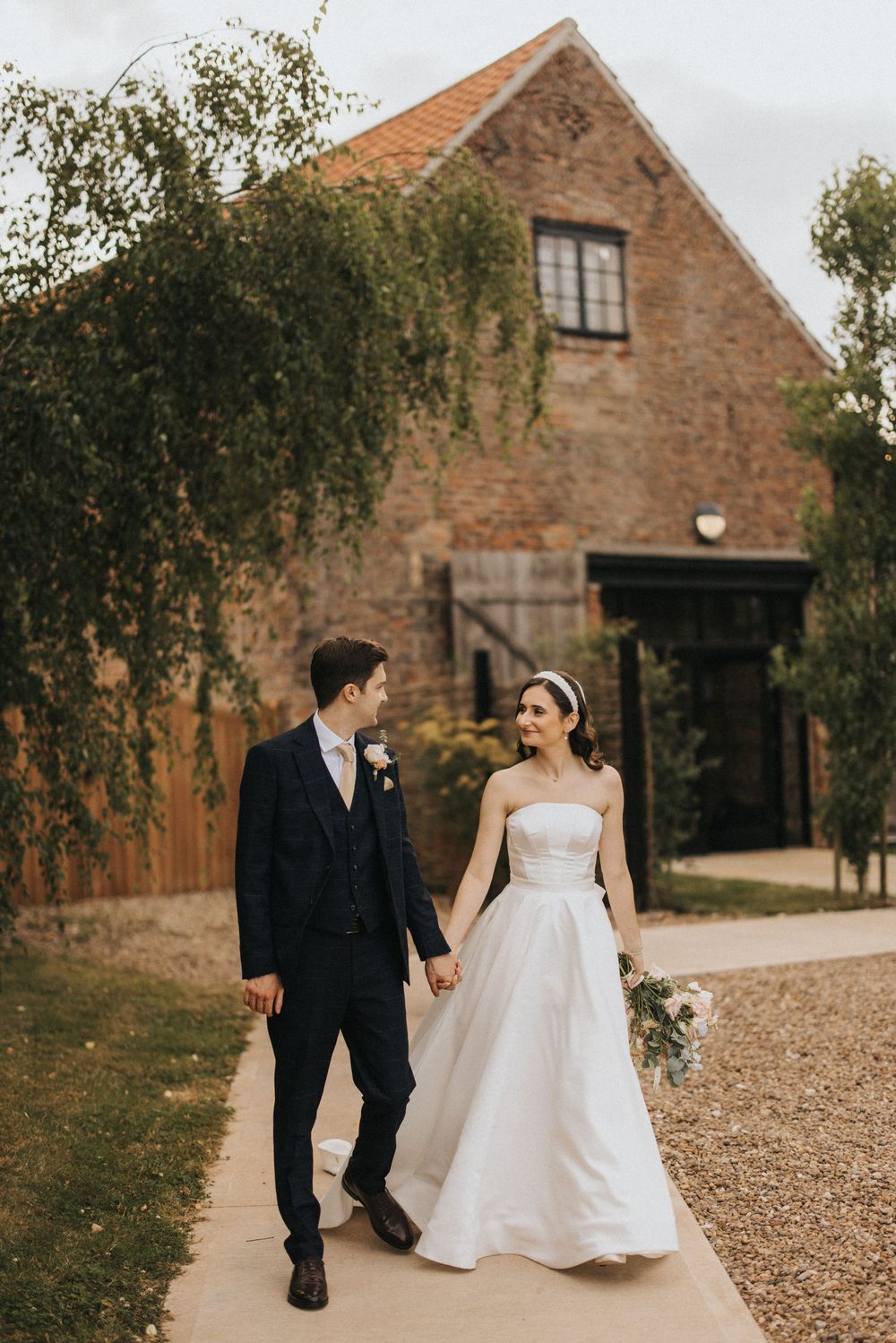 Bride and groom walk together along a rustic gravel path beside a stone barn building in autumn.