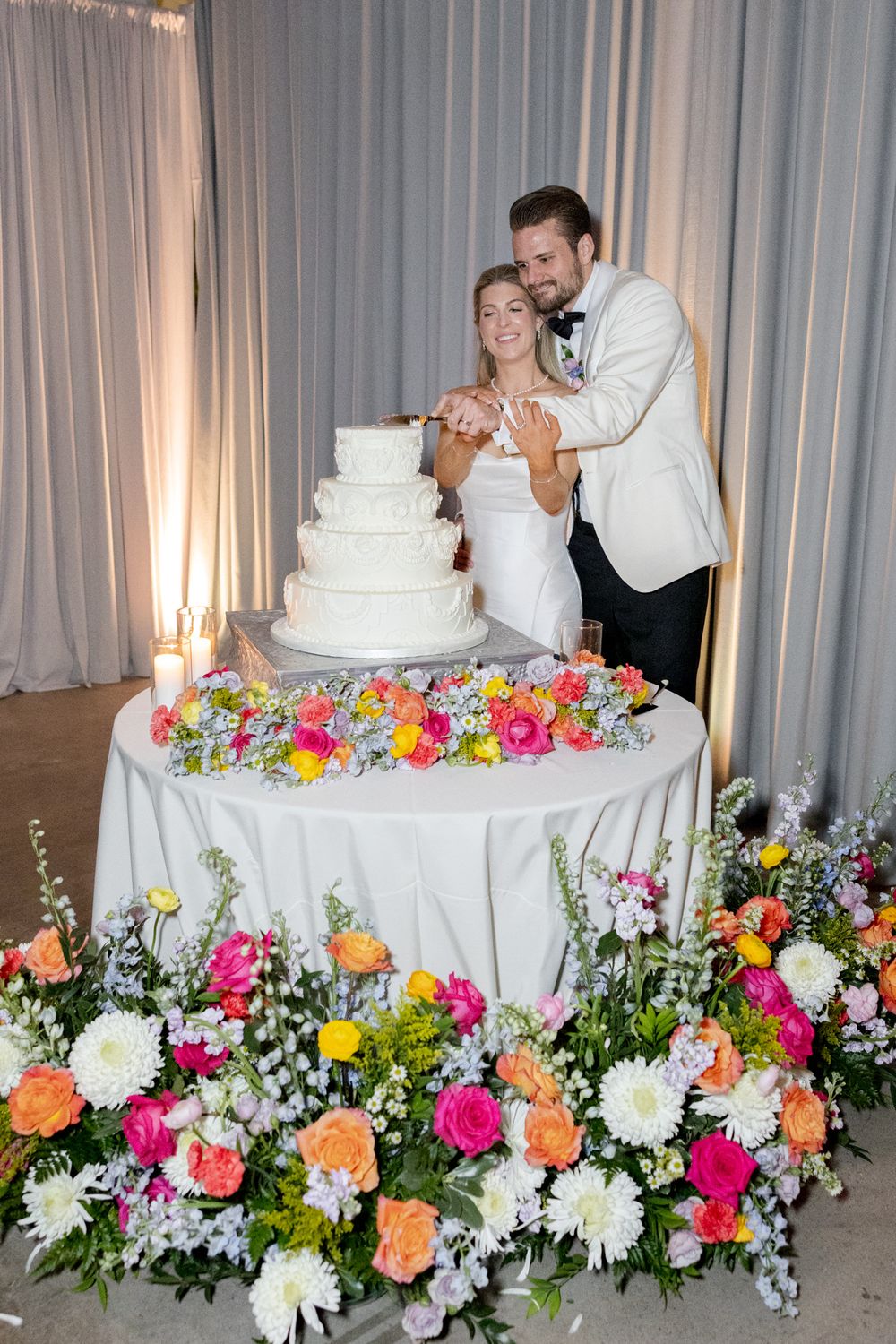Newlyweds cut their elegant white wedding cake surrounded by vibrant pink, orange and yellow floral arrangements.