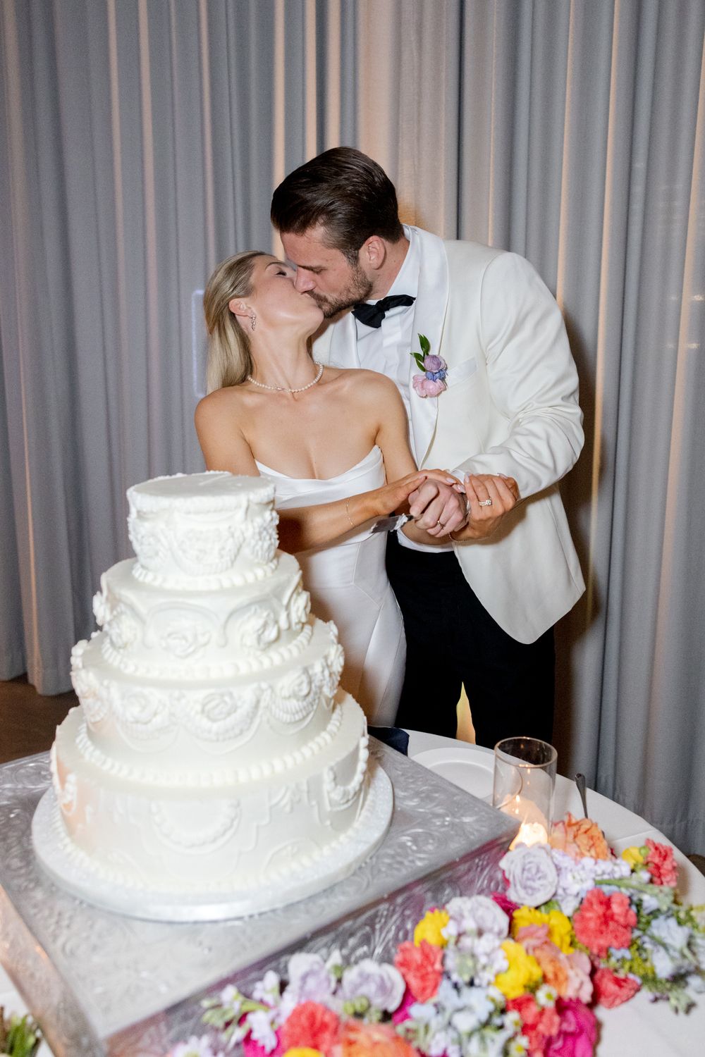 Romantic moment between newlyweds sharing a kiss while cutting their ornate white tiered wedding cake.