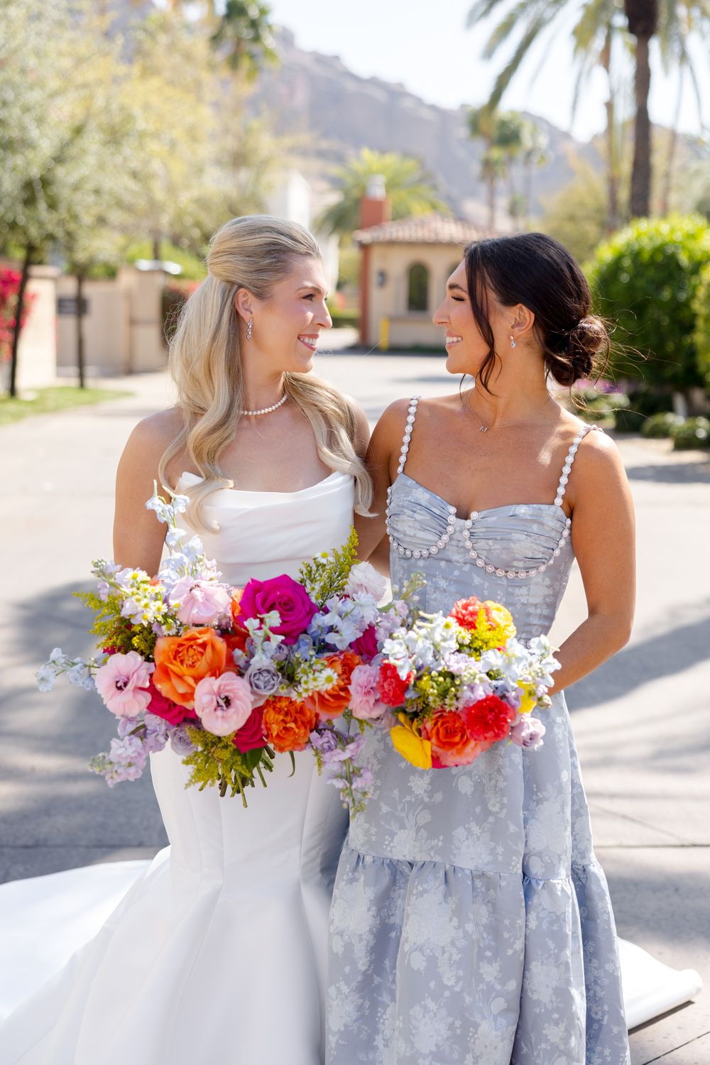 Bridesmaids in pale blue dresses carrying vibrant colorful bouquets pose outdoors with palm trees in background.