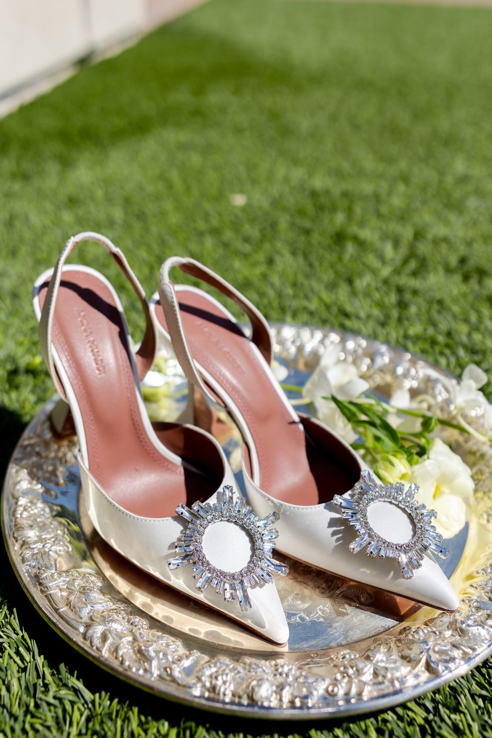 Elegant silver slingback bridal shoes with crystal sunburst ornaments displayed on a decorative metallic tray on grass.