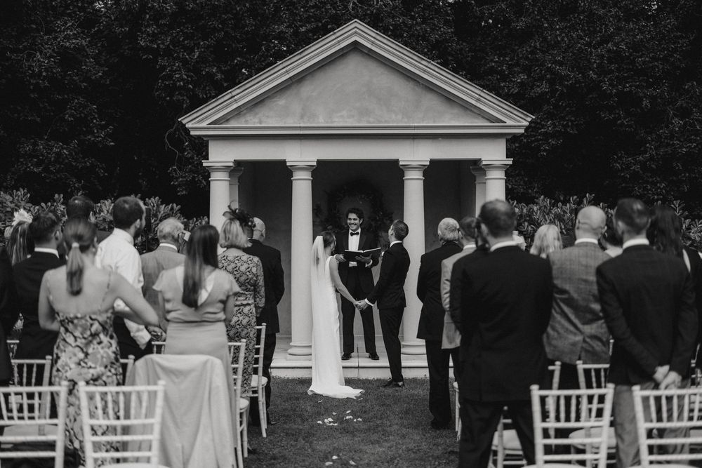 Black and white photo of an outdoor wedding ceremony at a classical white columned gazebo with guests seated in rows.