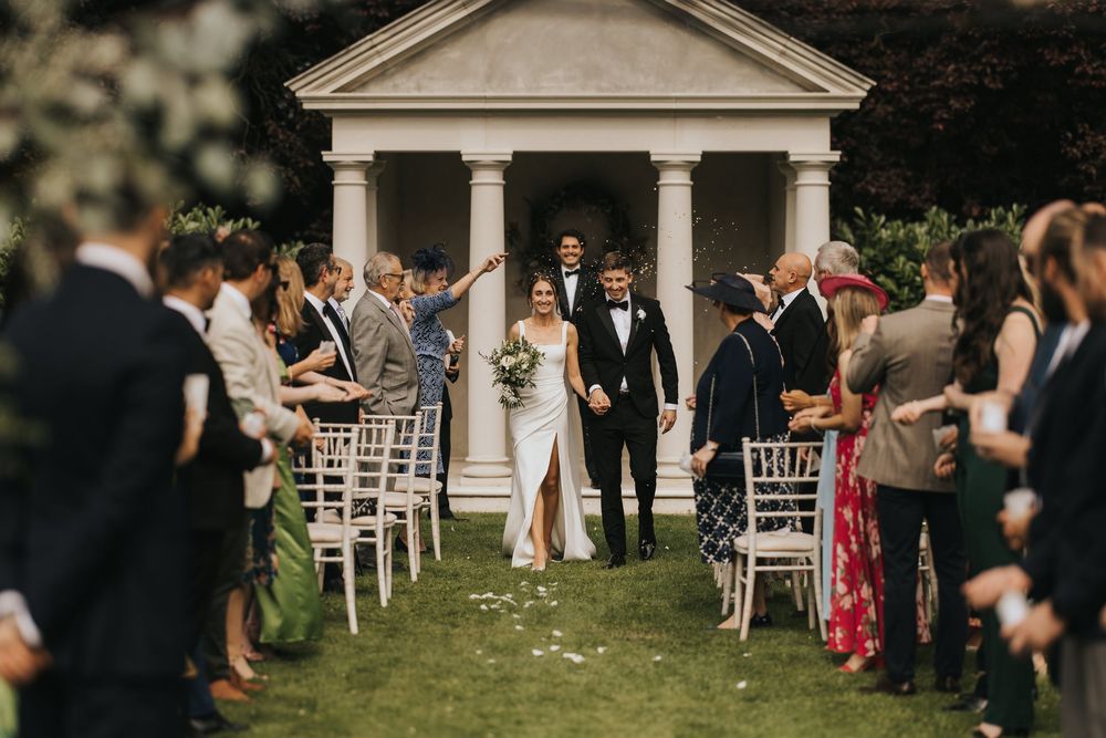 Wedding guests walk across a lush garden lawn in front of a white gazebo during an elegant outdoor ceremony.