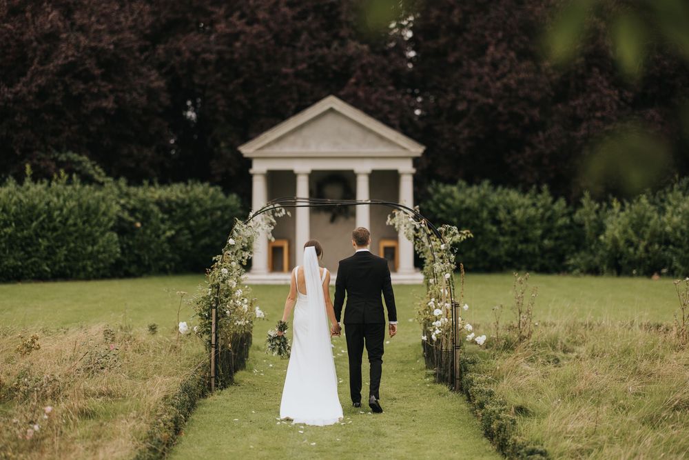 A couple walks down a garden path towards a white gazebo during an outdoor wedding ceremony.