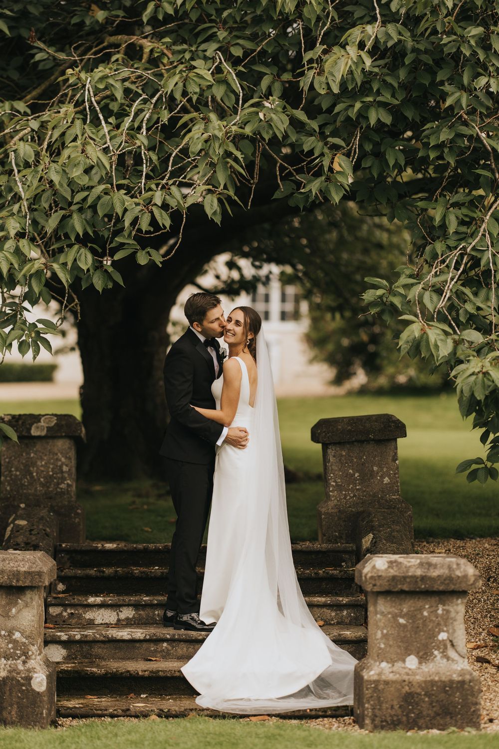 Elegant couple embraces on stone steps under a lush tree canopy in romantic garden wedding setting.