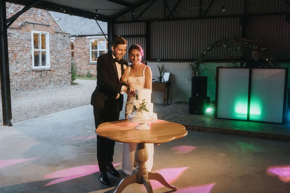 A couple cuts their wedding cake together in a rustic barn venue illuminated by colorful spotlights.