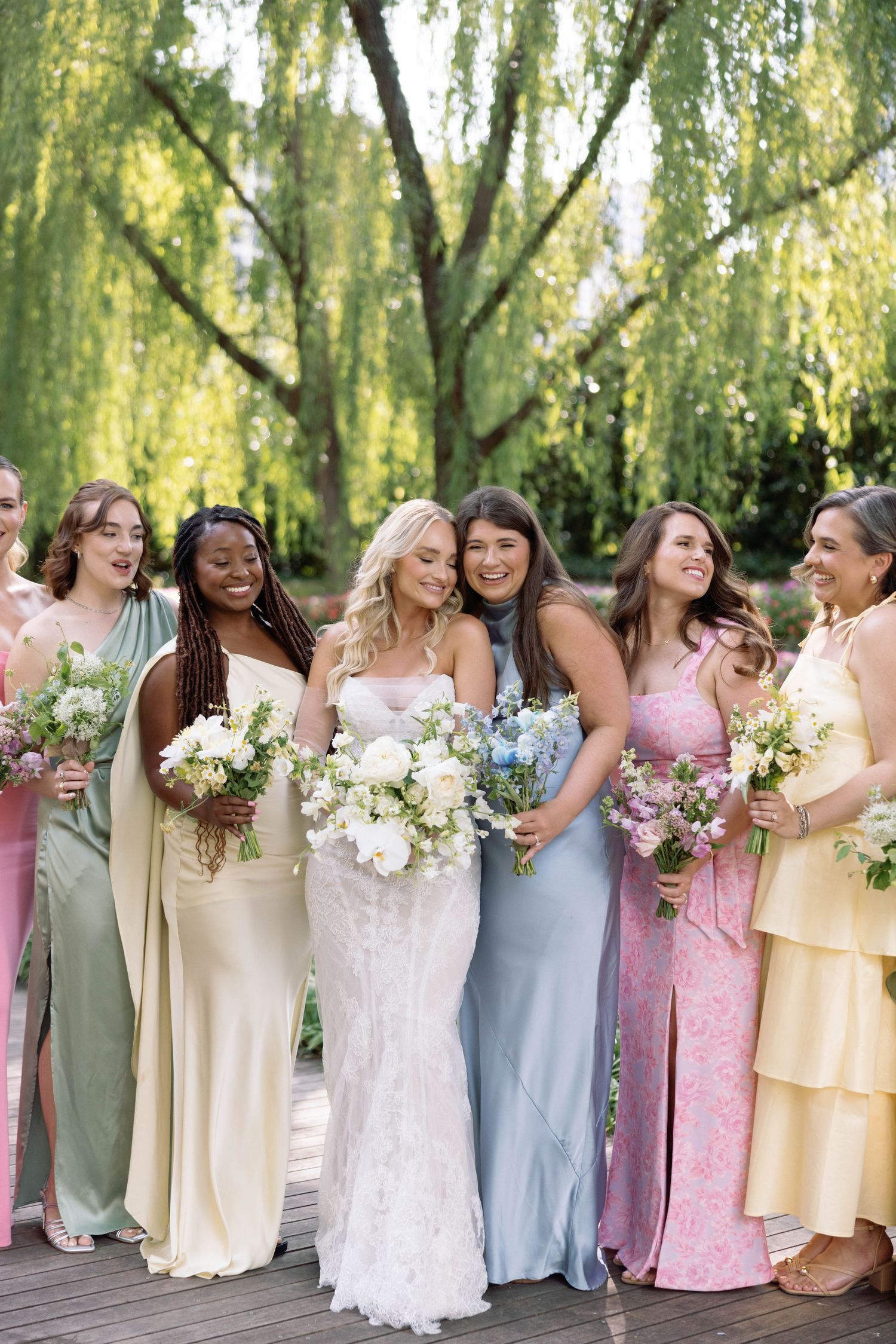 Bridal party in pastel dresses stands together on wooden deck under willow tree holding floral bouquets.