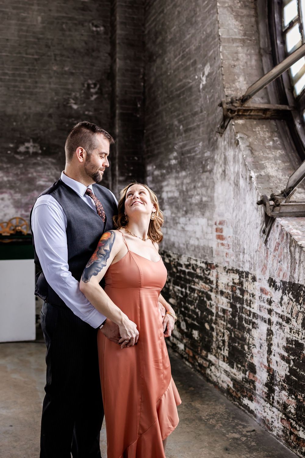 Couple embraces in warm intimate poses against weathered industrial brick backdrop.
