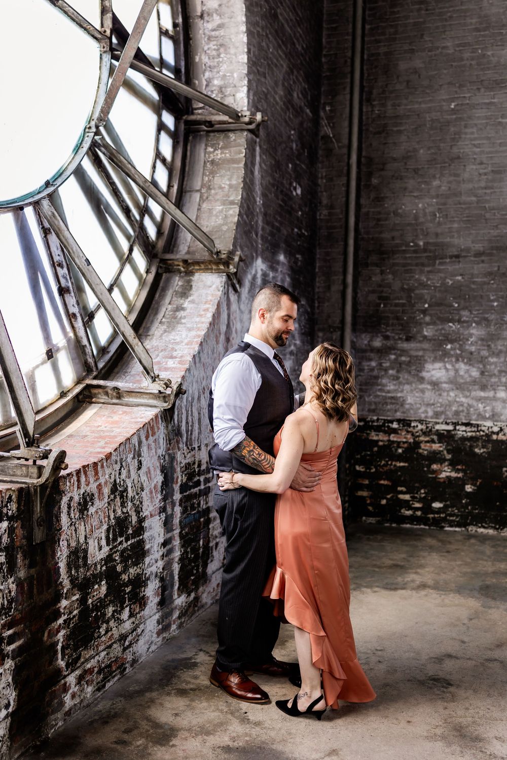 A couple dances in a romantic embrace near a large vintage clock face in an industrial brick building.